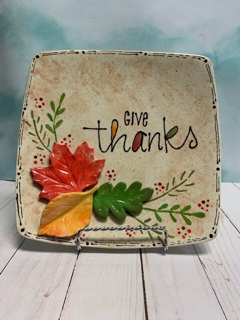 A square ceramic plate painted with colorful leaves with “give thanks” written in the middle