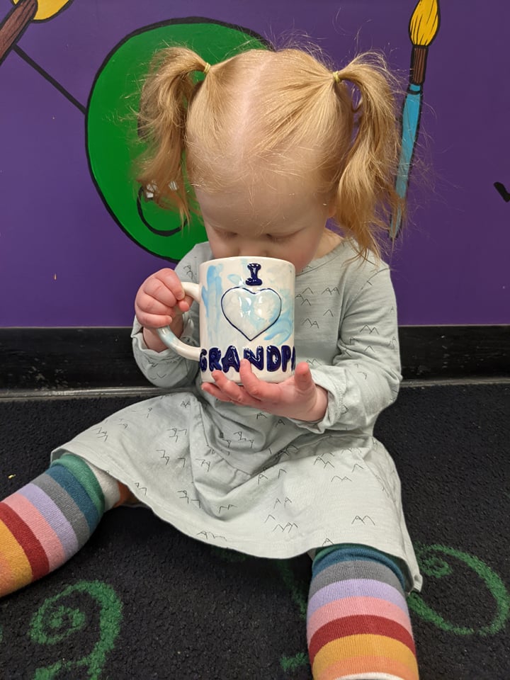 A toddler girl drinking from a mug on the floor.